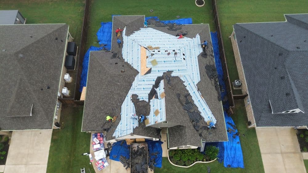 Aerial view of a house undergoing roof replacement with workers and blue tarps visible.
