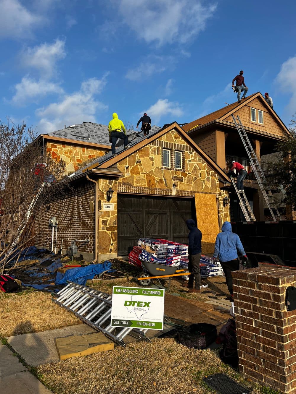 Workers repairing a roof on a stone house, ladders and materials visible, sunny day.