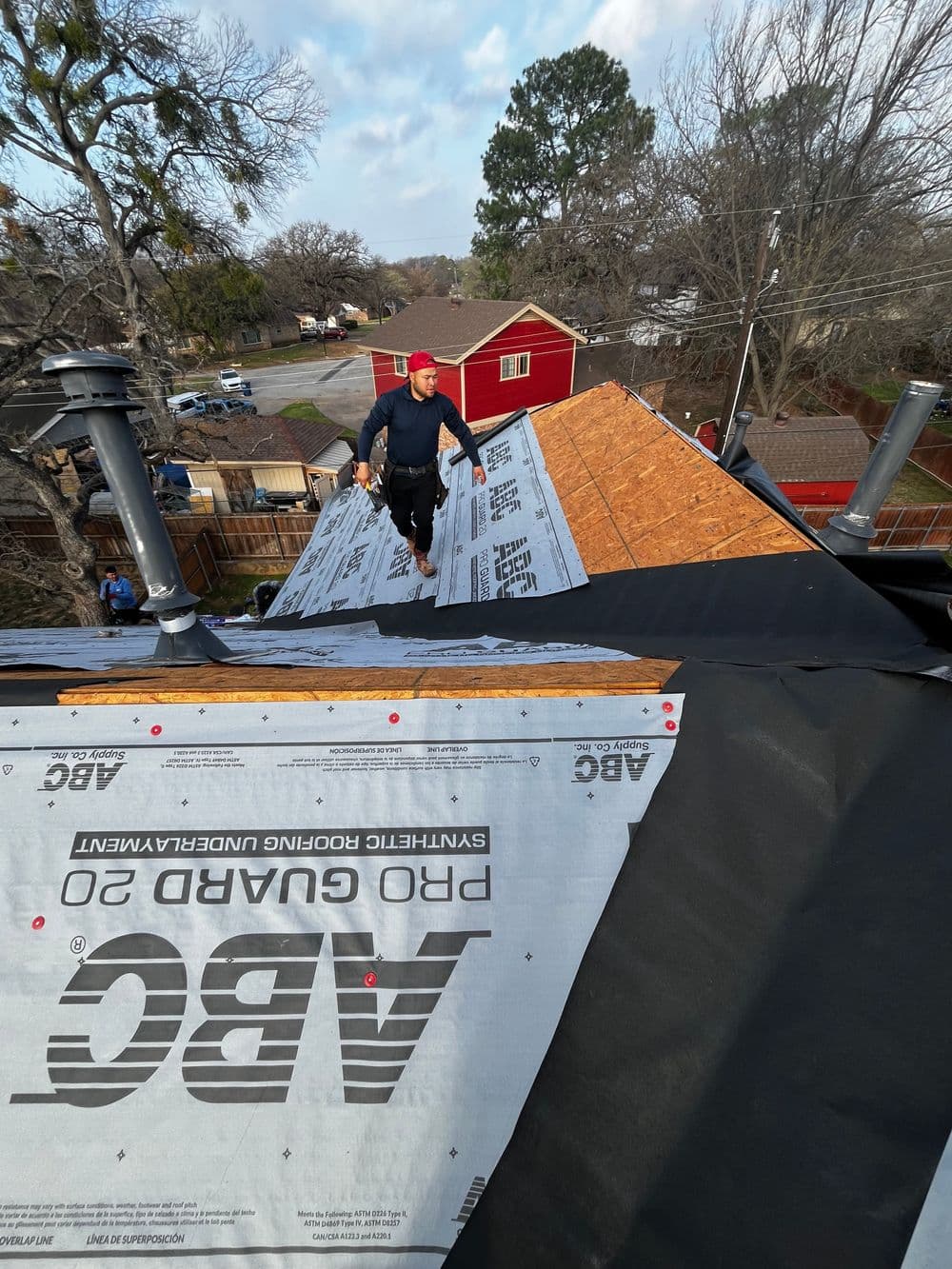 Worker installing roofing material on a sloped roof with trees and houses in the background.
